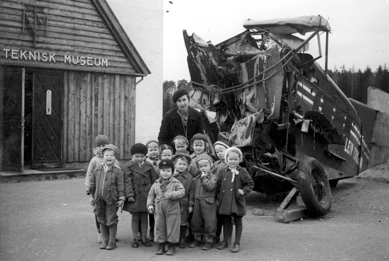 Ved inngangen til NTM: I forgrunnen barna og Fru Krag, i bakgrunnen et flyvrak