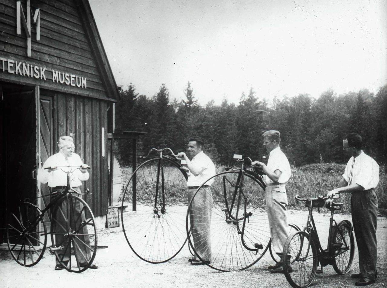 Fire menn, tre i hvite kortermede skjorter med hver sin sykkel. To av typen «velociped», også kalt «velte-petter»