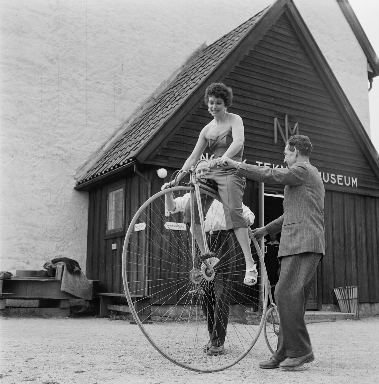 To menn støtter en kvinne på velociped. Velocipeden er høy, setehøyden er omtrent i skulderhøyde for mennene