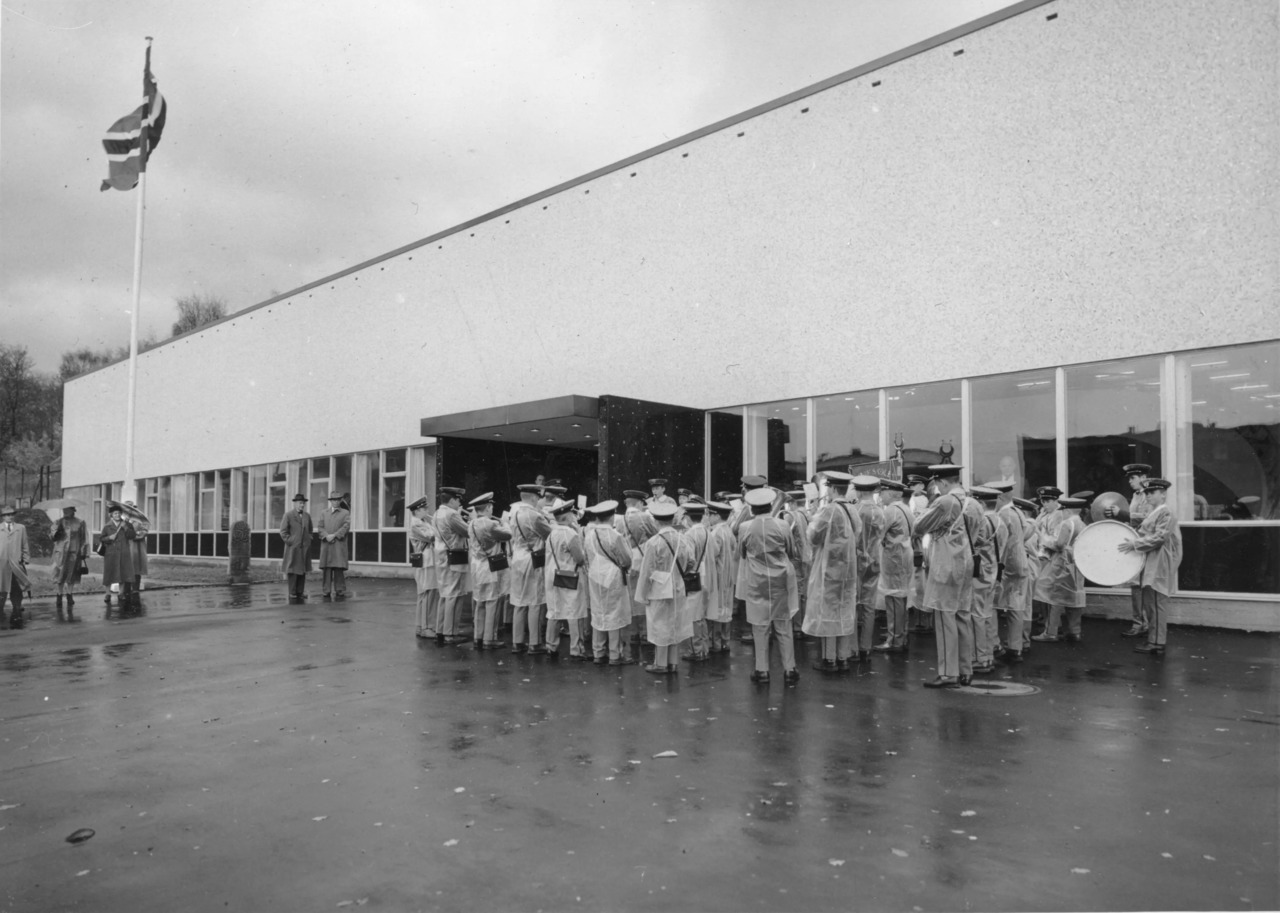 Korpsmedlemmer spiller ved inngangen. Det regner, og alle har regnfrakk over uniformen