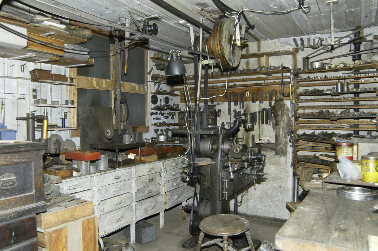 Verkstedet i kjelleren. Arbeidsbenker med skuffer, hyller med utstyr. Aksler og reimhjul i taket for drift av maskiner.